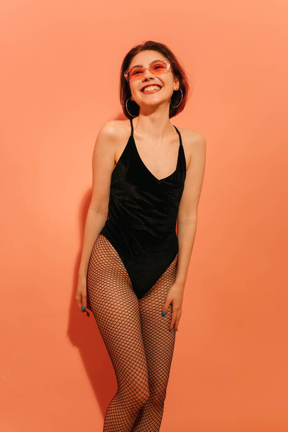 улыбающаяся молодая женщина позирует в черном телесном костюме и трико в сетке на оранжевом фоне - Фото, изображение