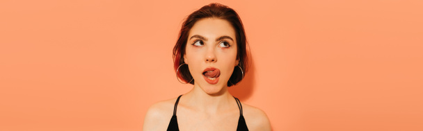 thoughtful young woman grimacing with sticking out tongue on orange background, banner - Photo, Image