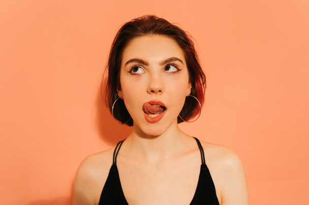 dreamy young woman grimacing with sticking out tongue on orange background - Фото, изображение