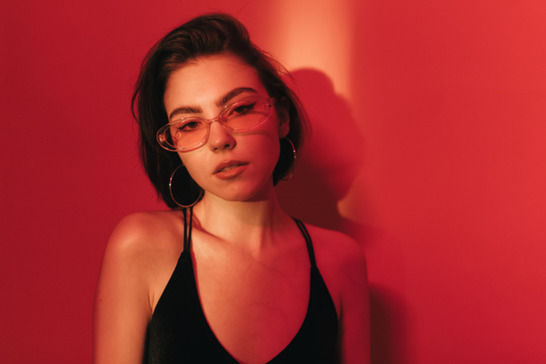 sensual woman with bob haircut in black bodysuit and sunglasses with half-opened mouth on red background with shadows - Фото, изображение