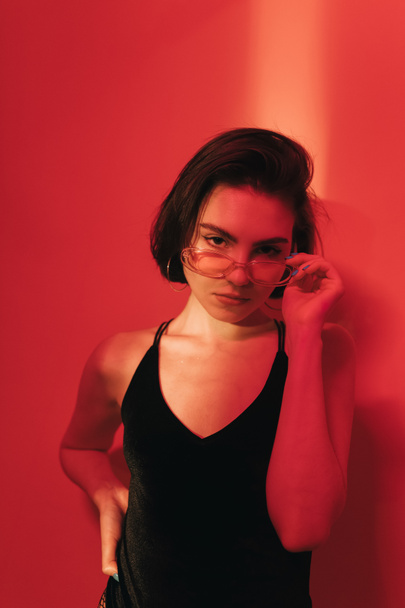 serious young woman with bob haircut in black bodysuit adjusting sunglasses and looking at camera on red background with shadows - Photo, image