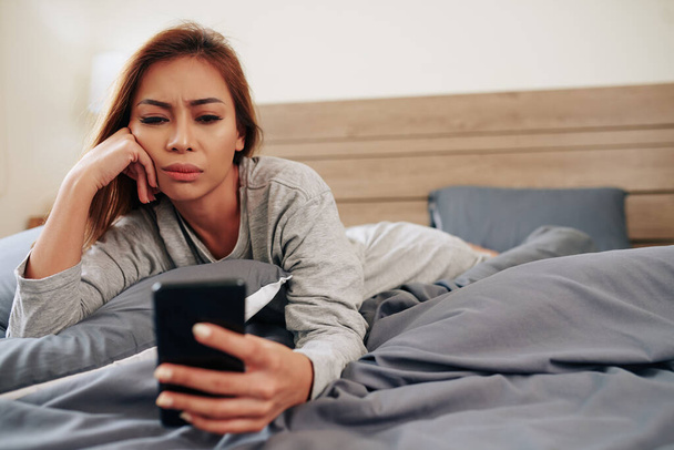 Triste mujer joven emocional leyendo malas noticias en la pantalla del teléfono inteligente al acostarse en la cama - Foto, Imagen