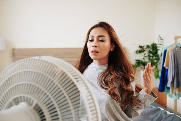Молода жінка стоїть перед вентилятором насолоджуючись свіжим повітрям в спекотний день
 - Фото, зображення