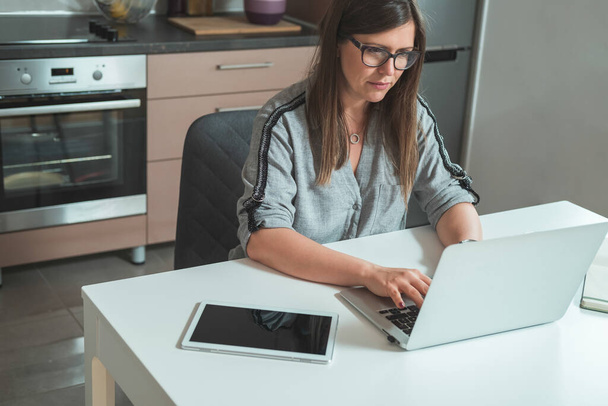 Piękna kobieta pracuje w domu Online na laptopie komputera.Poważny bizneswoman siedzi przy biurku i za pomocą laptopa do telekonferencji w godzinach porannych w kuchni. - Zdjęcie, obraz