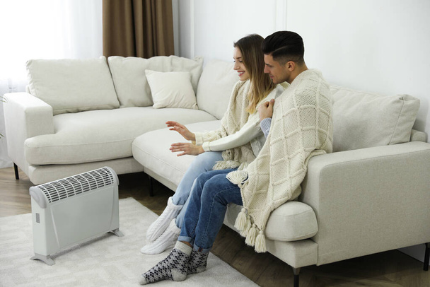 Happy couple sitting on sofa near electric heater at home - Фото, изображение