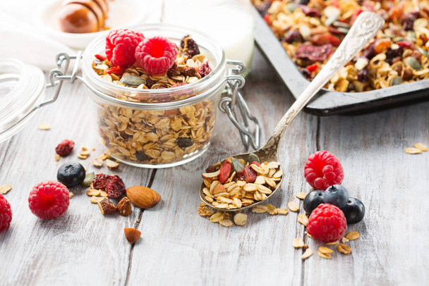 Fresh homemade granola. Handmade muesli with fresh berries, honey, seeds in glass jars on white wooden background. Selective focus, copy space. Healthy food concept. - 写真・画像