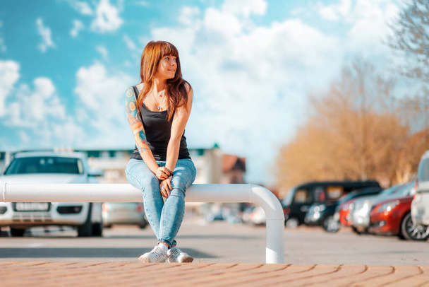 Eine junge lächelnde Frau mit tätowierter Hand sitzt auf der Leitplanke und blickt auf die rechte Seite. Draußen. - Foto, Bild