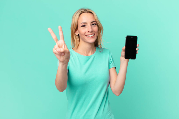 young blonde woman smiling and looking happy, gesturing victory or peace and holding a smart phone - Foto, Imagem