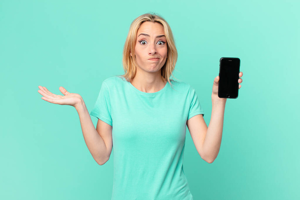 young blonde woman feeling puzzled and confused and doubting and holding a smart phone - Fotó, kép