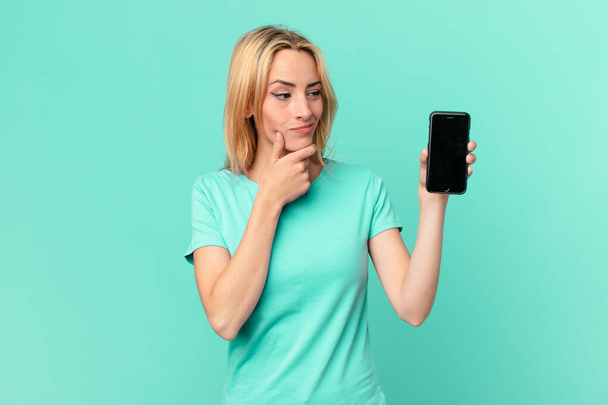 young blonde woman smiling with a happy, confident expression with hand on chin and holding a smart phone - Foto, Bild