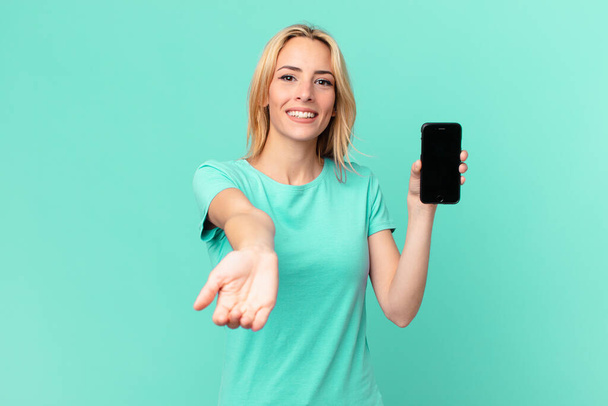 jeune femme blonde souriant joyeusement avec amical et offrant et montrant un concept et tenant un téléphone intelligent - Photo, image