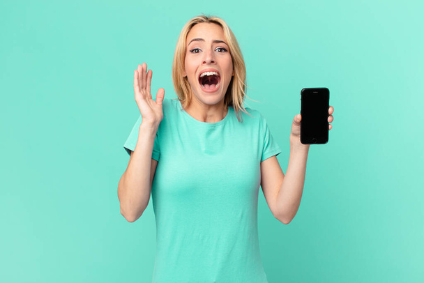 young blonde woman feeling happy and astonished at something unbelievable and holding a smart phone - Foto, Bild