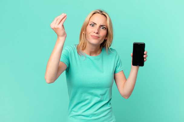 young blonde woman making capice or money gesture, telling you to pay and holding a smart phone - Photo, Image