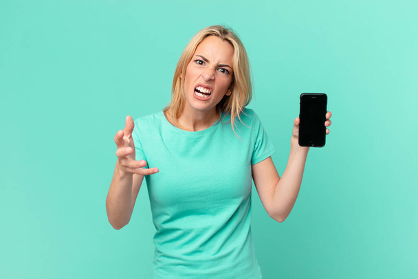 young blonde woman looking angry, annoyed and frustrated and holding a smart phone - Fotó, kép