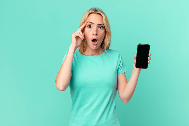 young blonde woman looking surprised, realizing a new thought, idea or concept and holding a smart phone - Фото, изображение