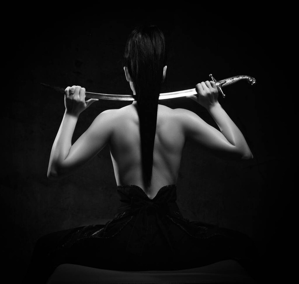 Black and white portrait of young pretty brunette woman sitting topless back to camera holding sword on shoulders - Fotoğraf, Görsel