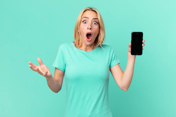 young blonde woman feeling extremely shocked and surprised and holding a smart phone - Zdjęcie, obraz