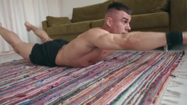 Молодой мускулистый мужчина тренируется дома и поднимает руки и ноги, лежа на коврике на животе. - Кадры, видео