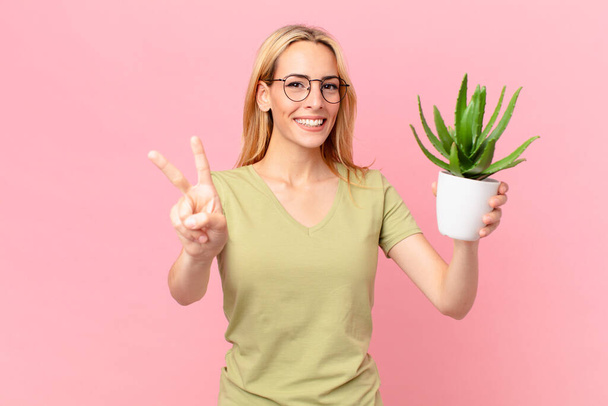 young blonde woman smiling and looking friendly, showing number two and holding a cactus - Photo, Image