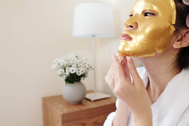Joven mujer asiática despegando rejuvenecedor e hidratante máscara de lámina dorada - Foto, imagen