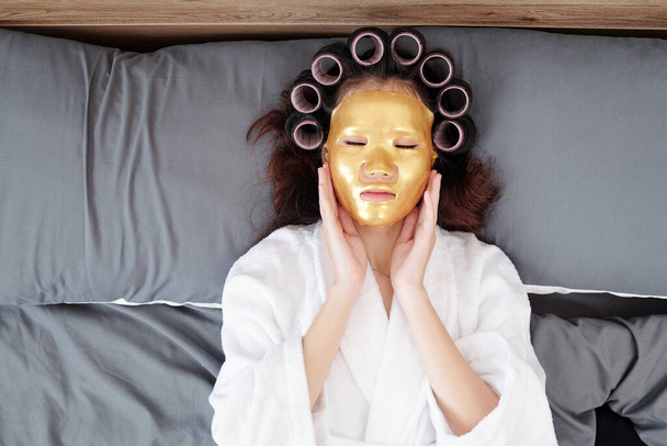 Молода азіатка відпочиває на ліжку з роликами для волосся і золотою маскою на обличчі. - Фото, зображення