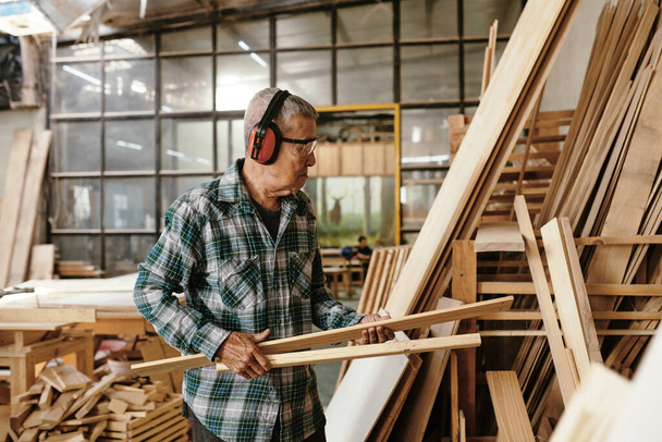 Carpinteiro sênior em earmuffs e camisa xadrez transportando pranchas de madeira para armazenamento de oficina - Foto, Imagem
