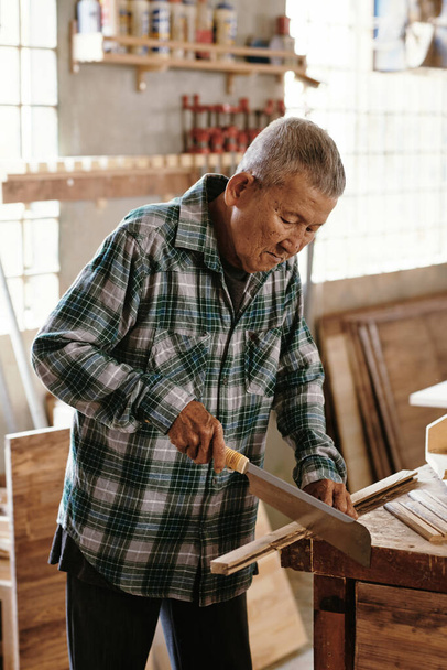 Senior menuisier découpe planche de bois avec scie japonaise ou nokogori à son établi - Photo, image