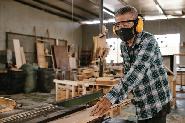 Senior man met beschermend masker en oorkappen werkend met hout in timmermanswerkplaats - Foto, afbeelding