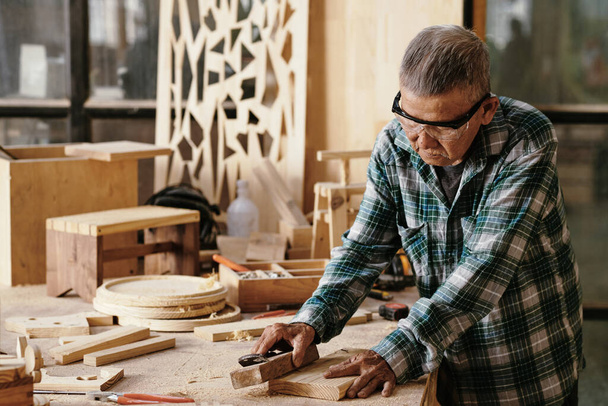 Carpinteiro sênior polimento placa pequena com bloco de madeira em sua bancada - Foto, Imagem