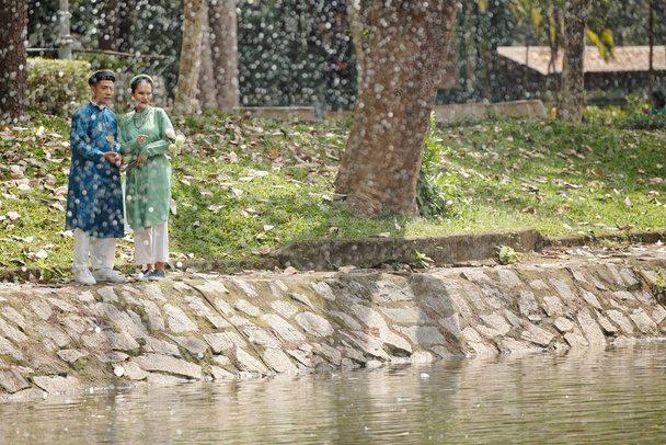 Just married young Vietnamese couple walking by pond in city park on rainy day - Photo, Image