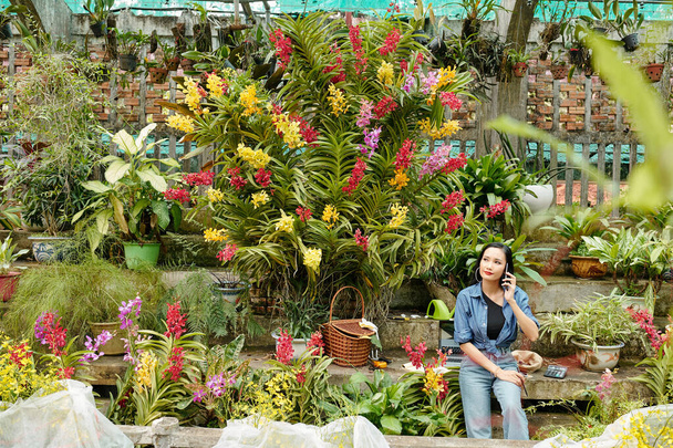 Bastante joven mujer asiática que trabaja en invernadero con muchas flores florecientes alrededor, ella hablando por teléfono con el cliente - Foto, Imagen