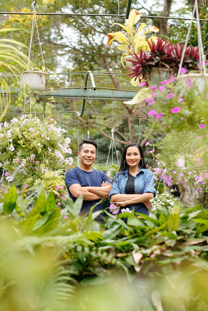 Feliz joven asiático propietarios de invernaderos de pie en invernadero con plantas y flores alrededor - Foto, Imagen