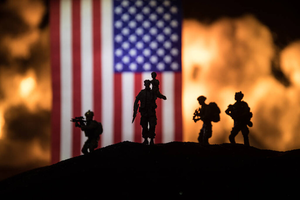 Amerykańska mała flaga na płonącym ciemnym tle. Koncepcja kryzysu wojny i konfliktów politycznych między narodami. Sylwetka uzbrojonego żołnierza przeciwko fladze USA. Skupienie selektywne - Zdjęcie, obraz