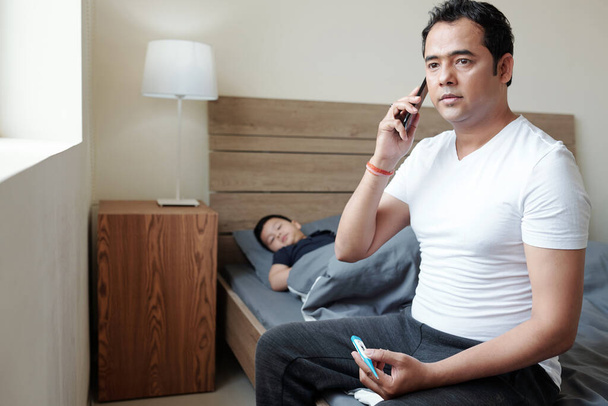 Hombre indio preocupado sentado en la cama de su hijo enfermo con fiebre y llamando al hospital - Foto, Imagen