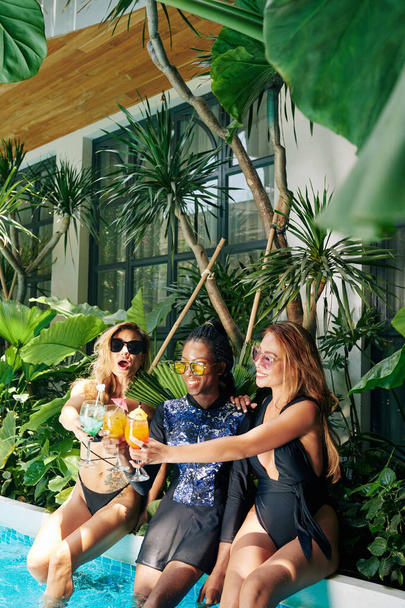 Vrolijke jonge vrouwelijke vrienden brengen tijd door bij het zwembad en klinkende glazen zoete fruit cocktails - Foto, afbeelding
