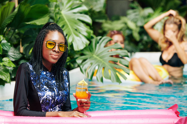Portret van lachende jonge zwarte vrouw in volledige dekking badpak en gele zonnebril staan in het zwembad en het drinken van non-alcoholische fruit cocktail - Foto, afbeelding