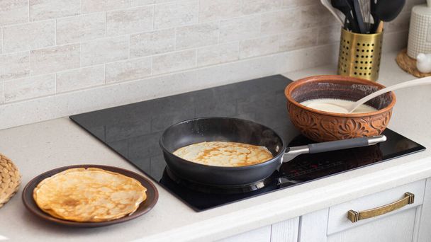 The process of making thin pancakes in a cozy home kitchen. Pancakes in a frying pan and in earthenware. Thin pancakes close-up. Light kitchen interior.  - Photo, image