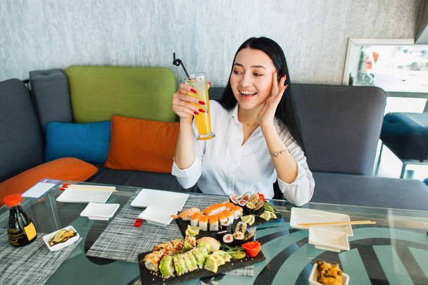 Young brunette Asian woman sitting in cafe at glass table, drinking fresh juice and enjoying her tasty lunch with sushi rolls at modern oriental cafe. - Foto, imagen
