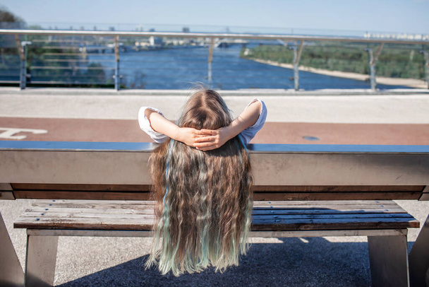 Morena chica preadolescente con reflejos de color azul mirando el río en el puente, vista trasera, día de verano - Foto, Imagen