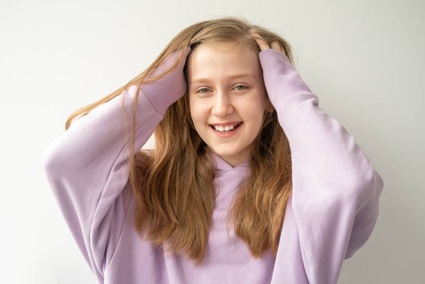 красивая девочка-подросток с длинными волосами в фиолетовом мундире, прикасающаяся к своим волосам, стоящим у белой стены. - Фото, изображение