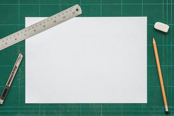 Old green dirty cutting mat, with empty white paper A4 size, stainless steel ruler, cutter knife, pencil and eraser, top view of workspace, copy space for text and graphic. - Photo, Image