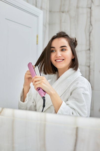 woman using a hair straightener in bathroom. - Foto, afbeelding