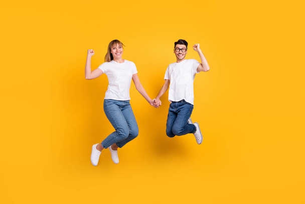 Full length photo of adorable sweet persons dressed white t-shirt jumping holding hands rising fists isolated yellow color background - Fotó, kép