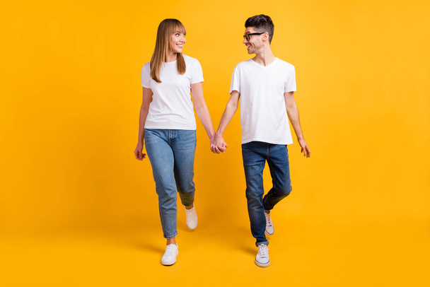 Full length photo of sweet charming couple wear t-shirt spectacles holding hands look each other isolated yellow color background - Φωτογραφία, εικόνα
