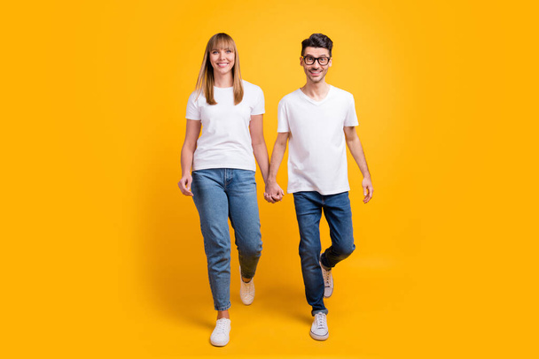 Full length photo of funky cute two persons dressed white t-shirt walking holding hands arms isolated yellow color background - Fotó, kép