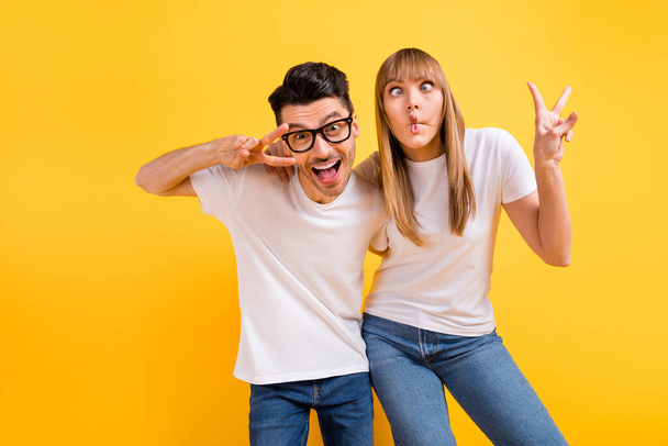 Photo of charming pretty young couple wear white t-shirt spectacles dancing showing v-signs isolated yellow color background - Foto, Imagen
