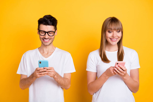 Photo of cute charming two persons dressed white t-shirt glasses typing modern devices isolated yellow color background - Foto, afbeelding