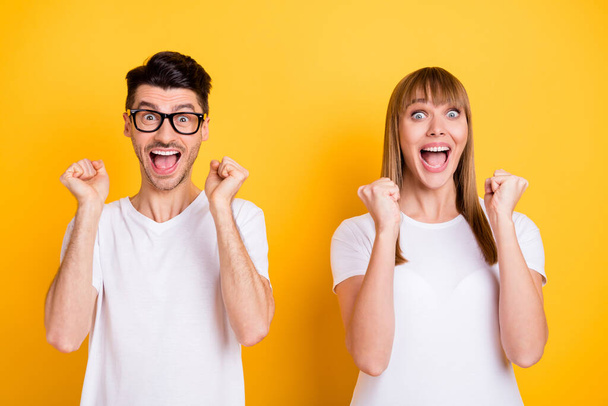 Photo of funny lucky young couple wear white t-shirt spectacles rising fists isolated yellow color background - Fotoğraf, Görsel