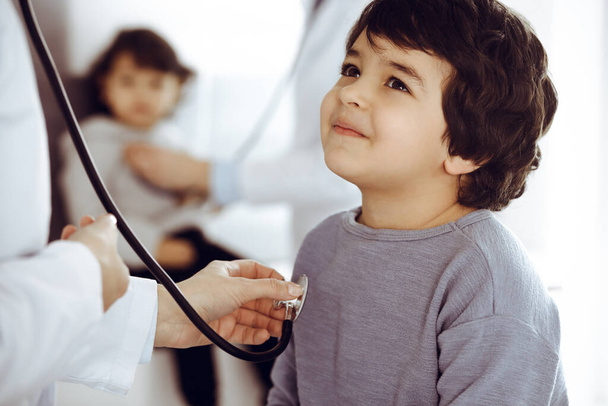 Doctor-woman examining a child patient by stethoscope. Cute arab boy and his brother at physician appointment. Medicine concept - Photo, Image