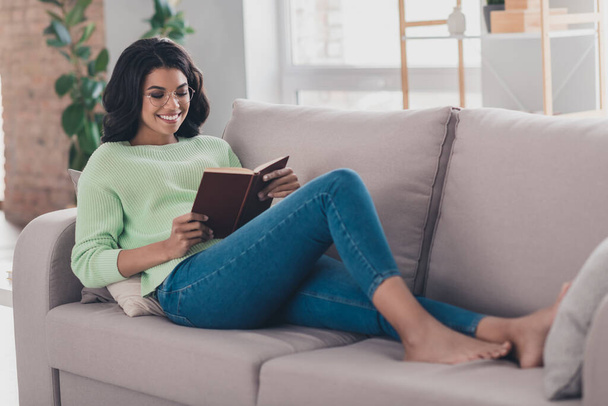 Full size profile photo of charming dark skin girl lying on couch hold book toothy smile weekend home indoors - Photo, image
